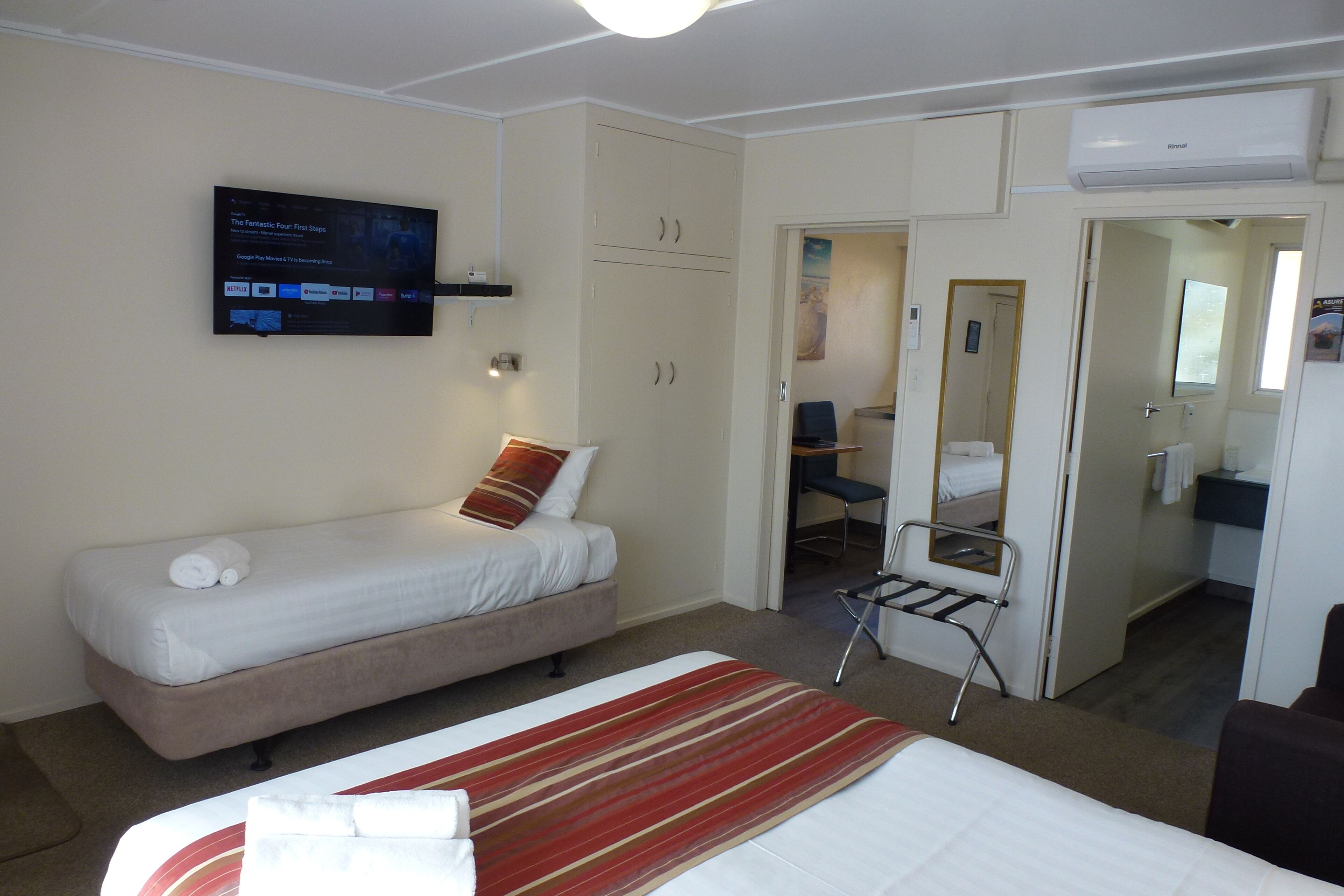 A hotel room with a single bed, a TV mounted on the wall, a wardrobe, and a bathroom visible through an open door.