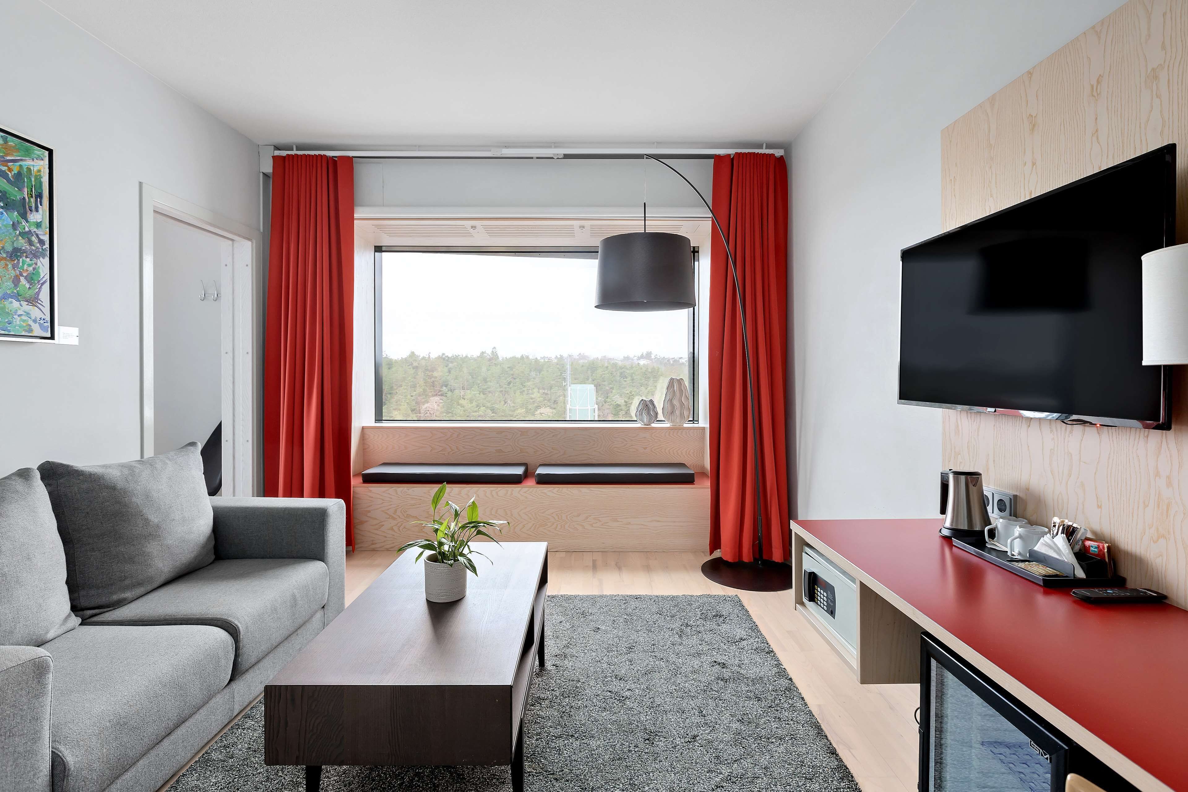 A modern living room with a grey sofa, a wooden coffee table, a red accent wall, a flat-screen TV, and a large window with red curtains.