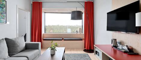 A modern living room with a grey sofa, a wooden coffee table, a red accent wall, a flat-screen TV, and a large window with red curtains.