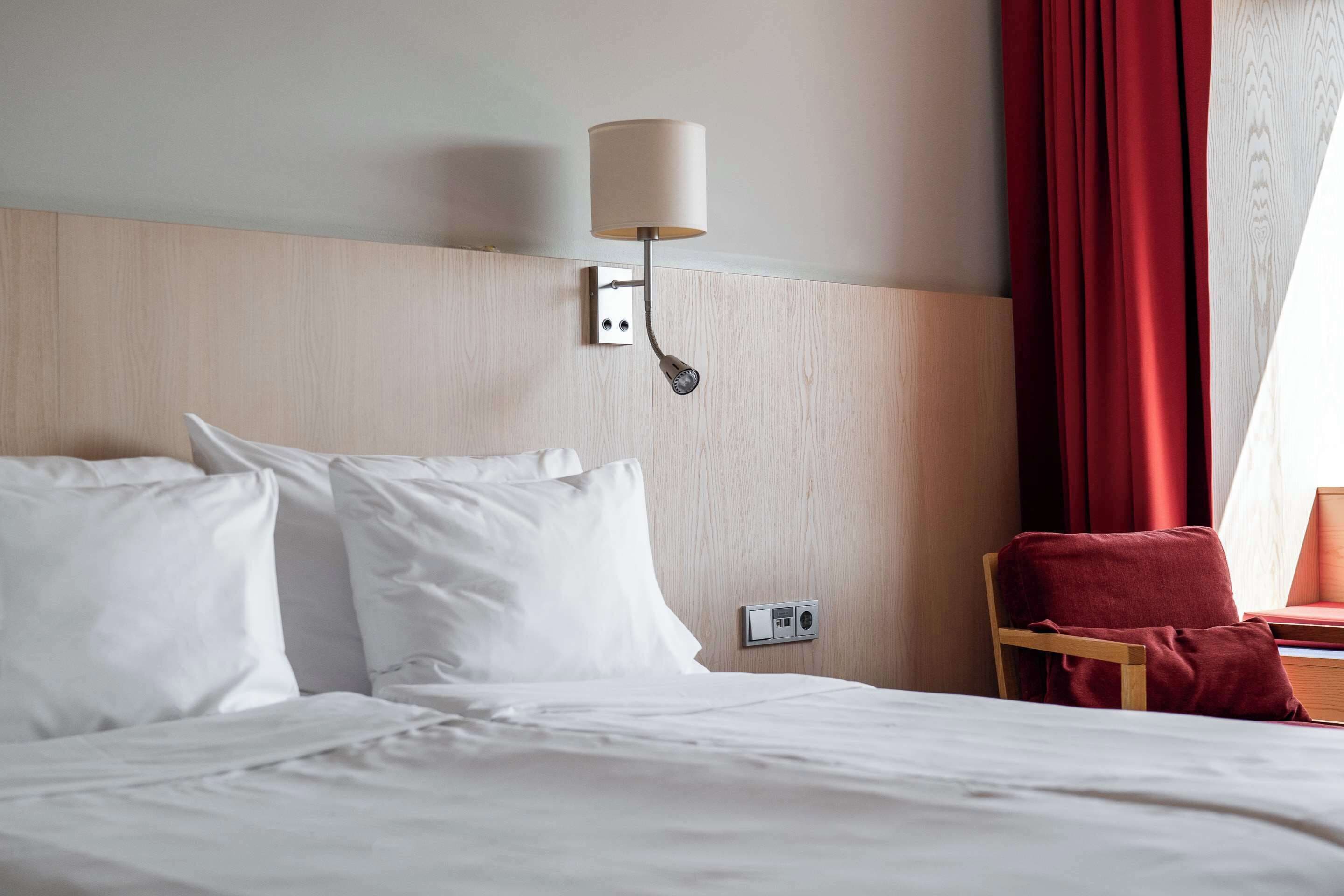 A hotel room with a bed, a red chair, a wall-mounted lamp, and a power outlet.