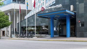 Entrada de un hotel moderno con un toldo azul, banderas y bicicletas estacionadas afuera.