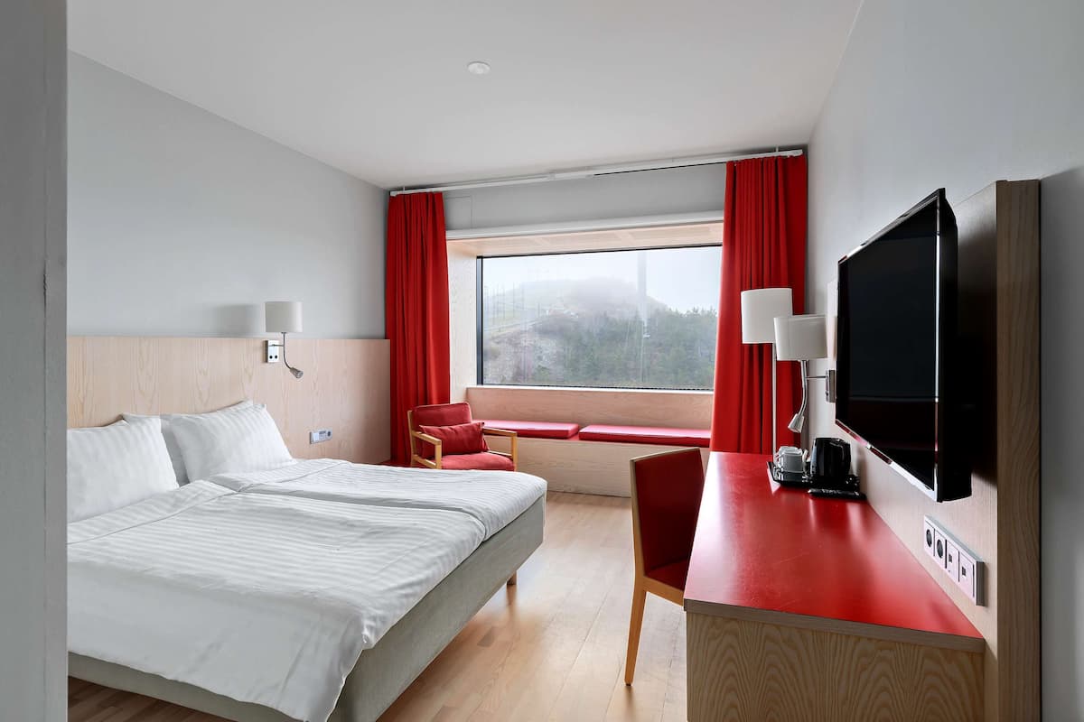 Une chambre d’hôtel avec un lit, un bureau avec une télévision, une chaise et une fenêtre avec des rideaux rouges.