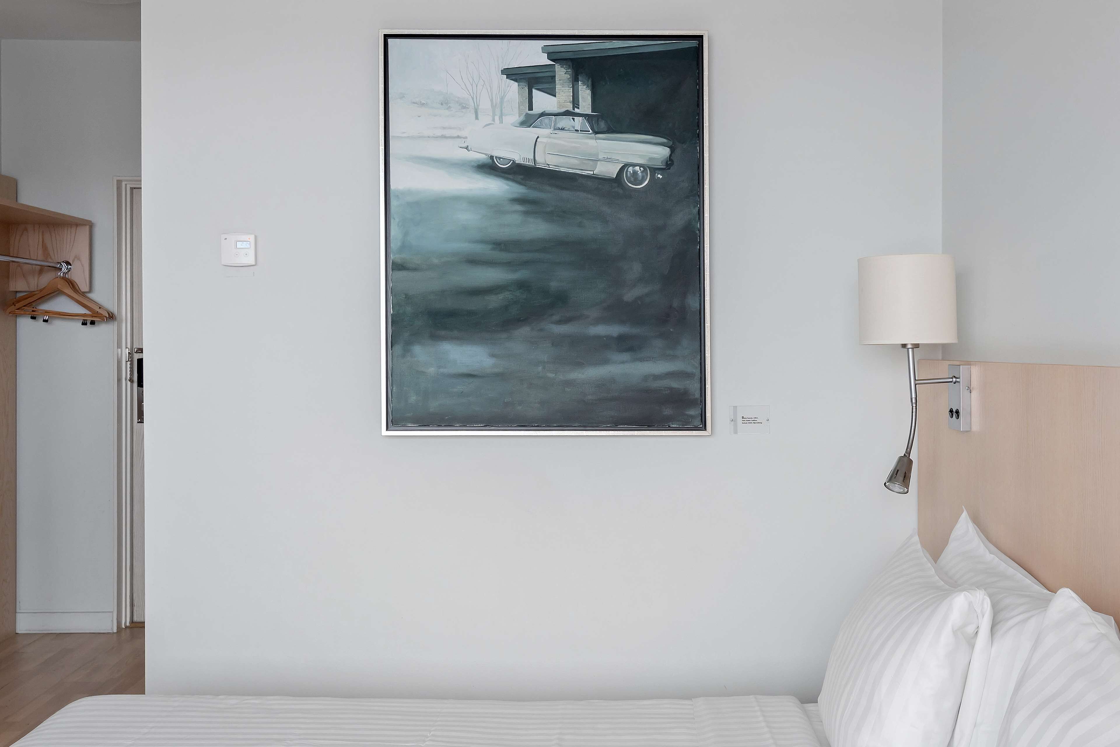 A bedroom with a bed, a wall-mounted lamp, and a framed artwork of a car.
