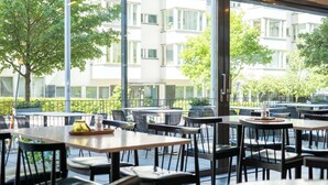 Un espacio exterior para comer con mesas y sillas, ventanas amplias y vistas a un edificio y árboles.