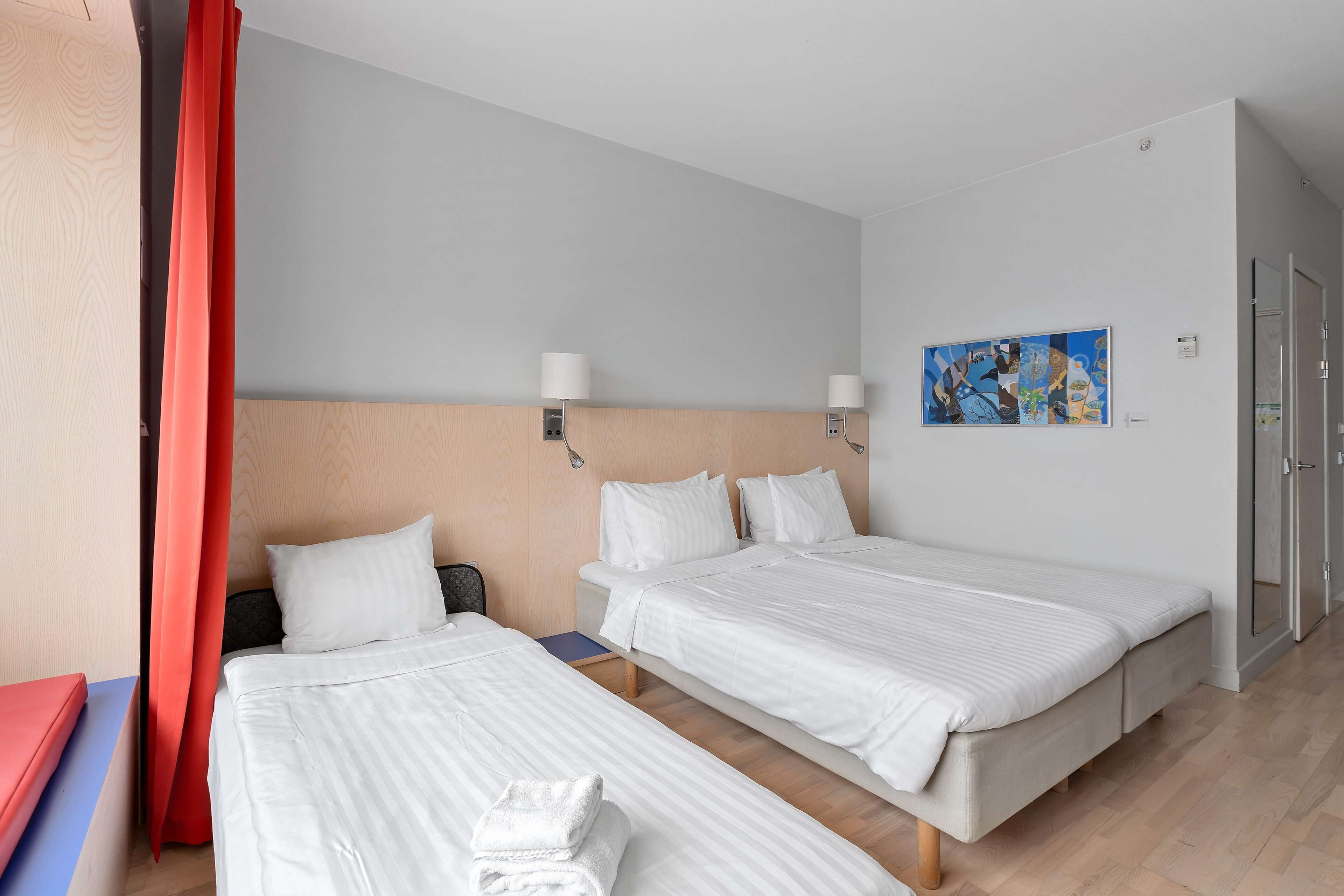 Habitación de hotel con dos camas, cortina roja, cabecero de madera, un cuadro en la pared y suelo de madera.