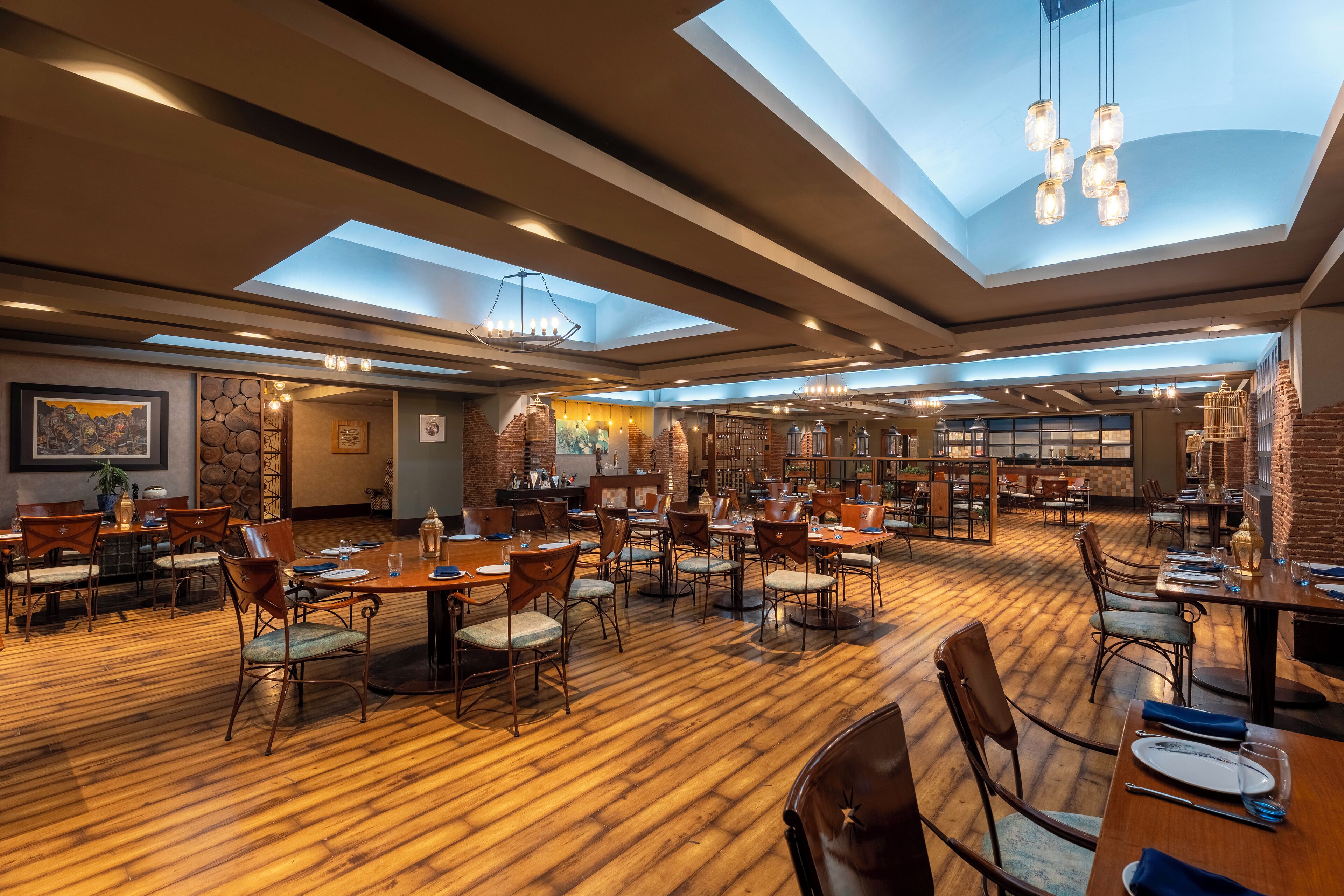 A spacious dining area with multiple tables and chairs, a skylight ceiling, and a brick wall.