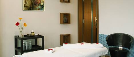 A massage room with a massage table, a chair, a shelf with bottles, and a painting on the wall.