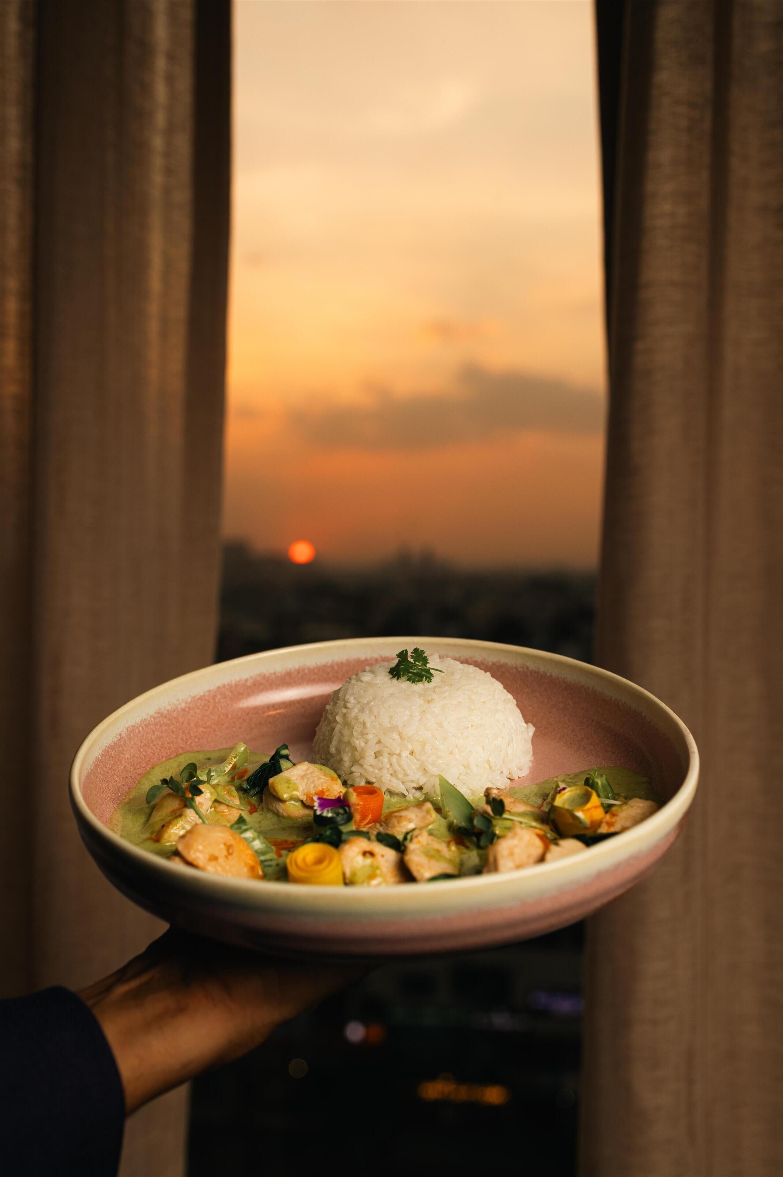 Un tazón de arroz y vegetales con vista al atardecer a través de cortinas.