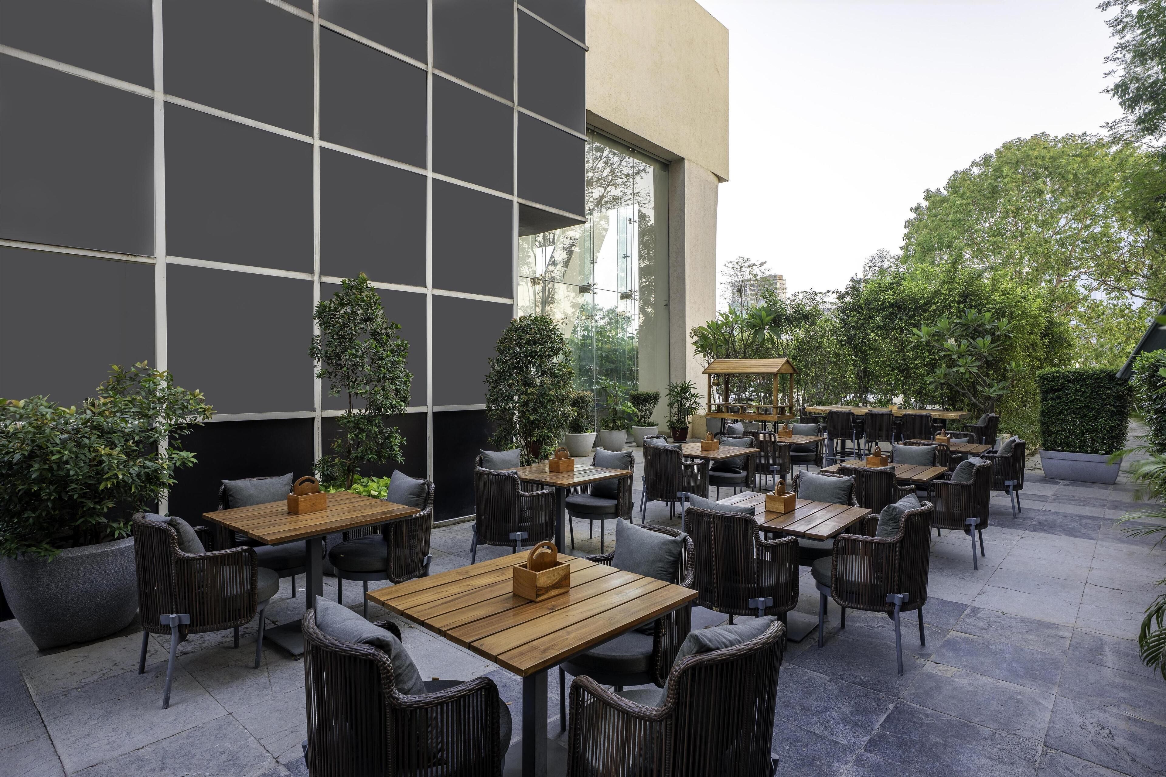 An outdoor patio area with multiple tables and chairs, surrounded by potted plants and a modern building facade.