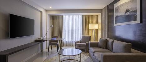 A modern hotel room with a flat-screen TV, a sofa, a coffee table, a desk with a lamp, and a large window with curtains.