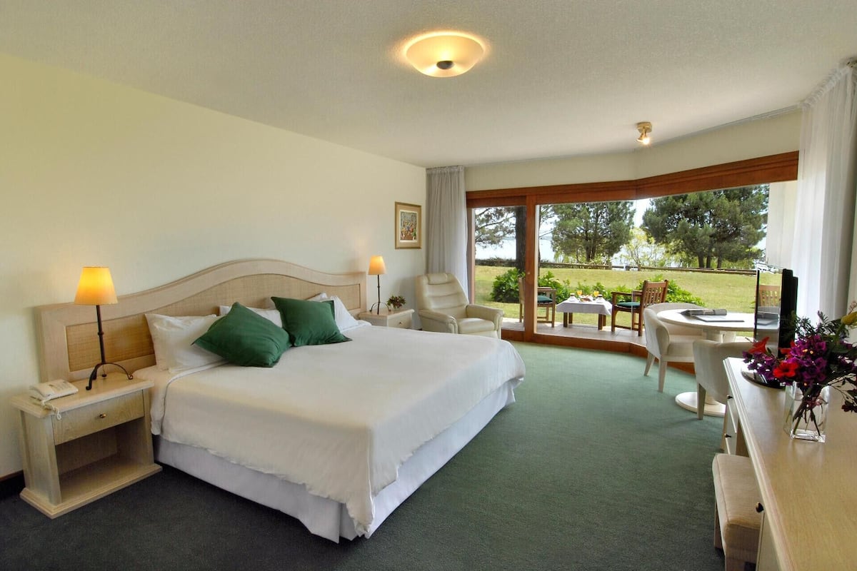 A hotel room with a bed, two bedside lamps, a desk with a chair, a small table with a chair, and a view of the outdoors.