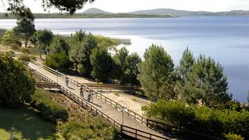 Pasarela de madera a lo largo de un lago con ciclistas, rodeada de vegetación y colinas al fondo.