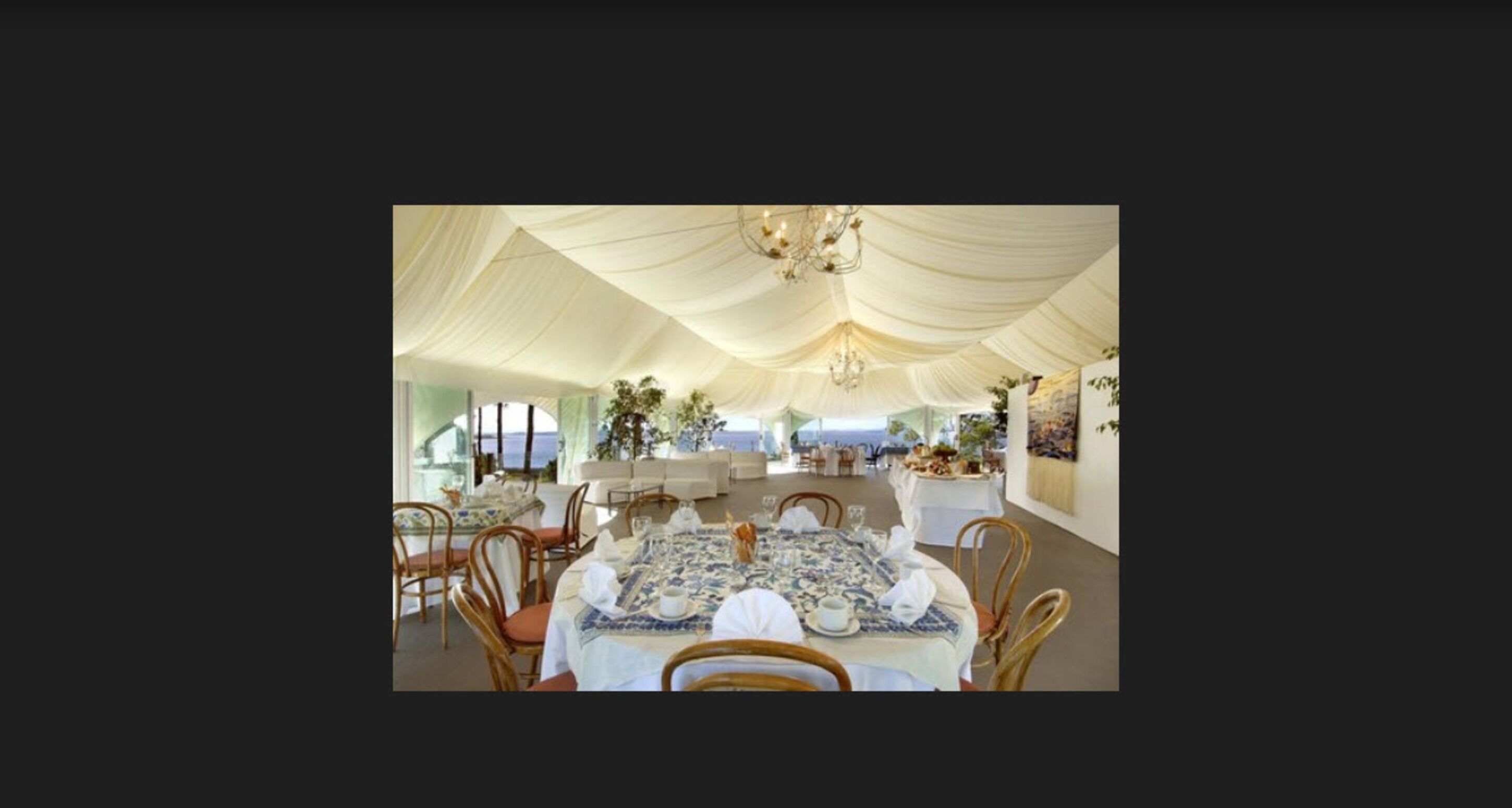 An indoor dining area with tables set for a meal, a chandelier, and a view of the sea.