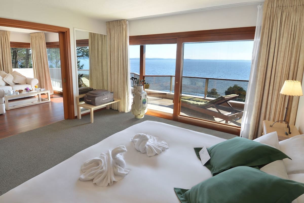 A hotel room with a large bed, a seating area with a view of the sea, and a balcony with a table and chairs.