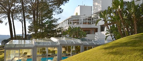 A modern house with a glass-enclosed pool area, surrounded by lush greenery and a clear view of the ocean.