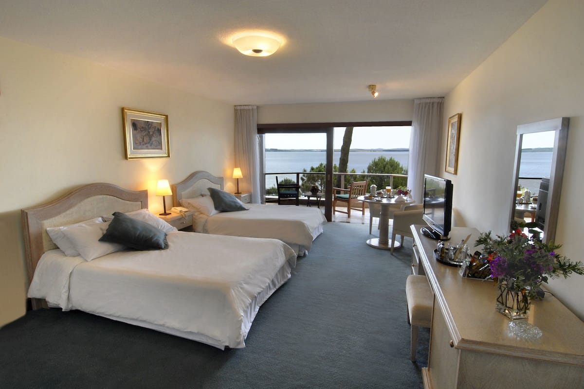 A hotel room with two beds, a TV, a desk, and a balcony with a view of the sea.