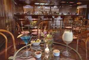 Zona de bar con sillas de madera, una mesa redonda con una vela y varios botellas y vasos en los estantes.