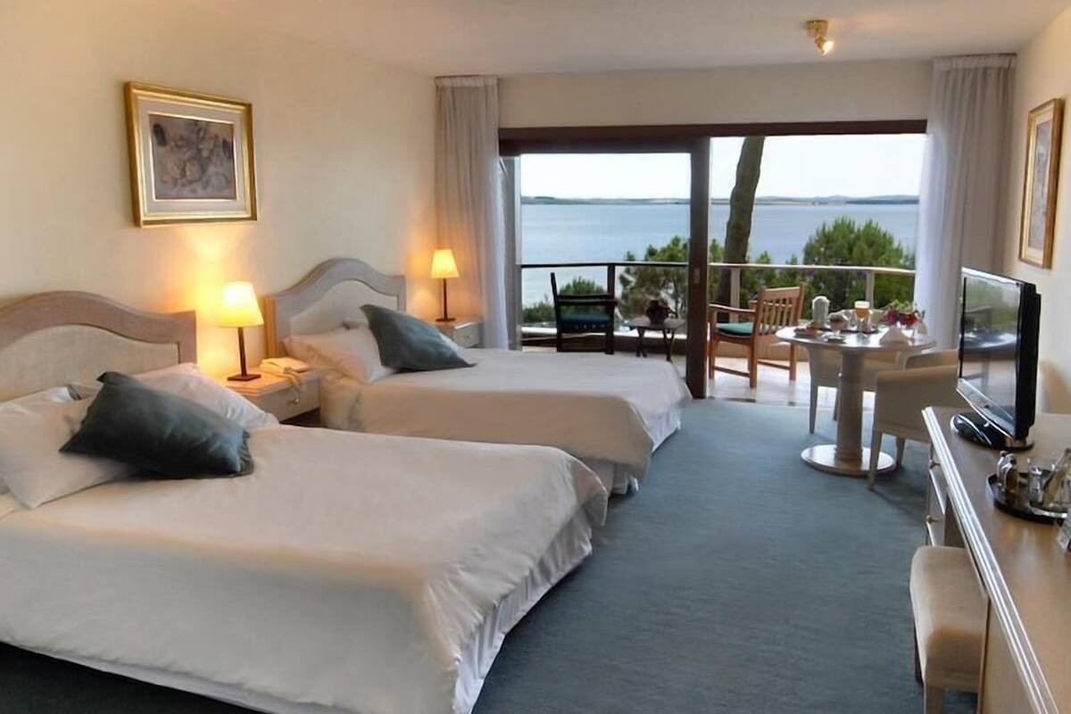 A hotel room with two beds, a balcony with a table and chairs, a TV, and a painting on the wall.