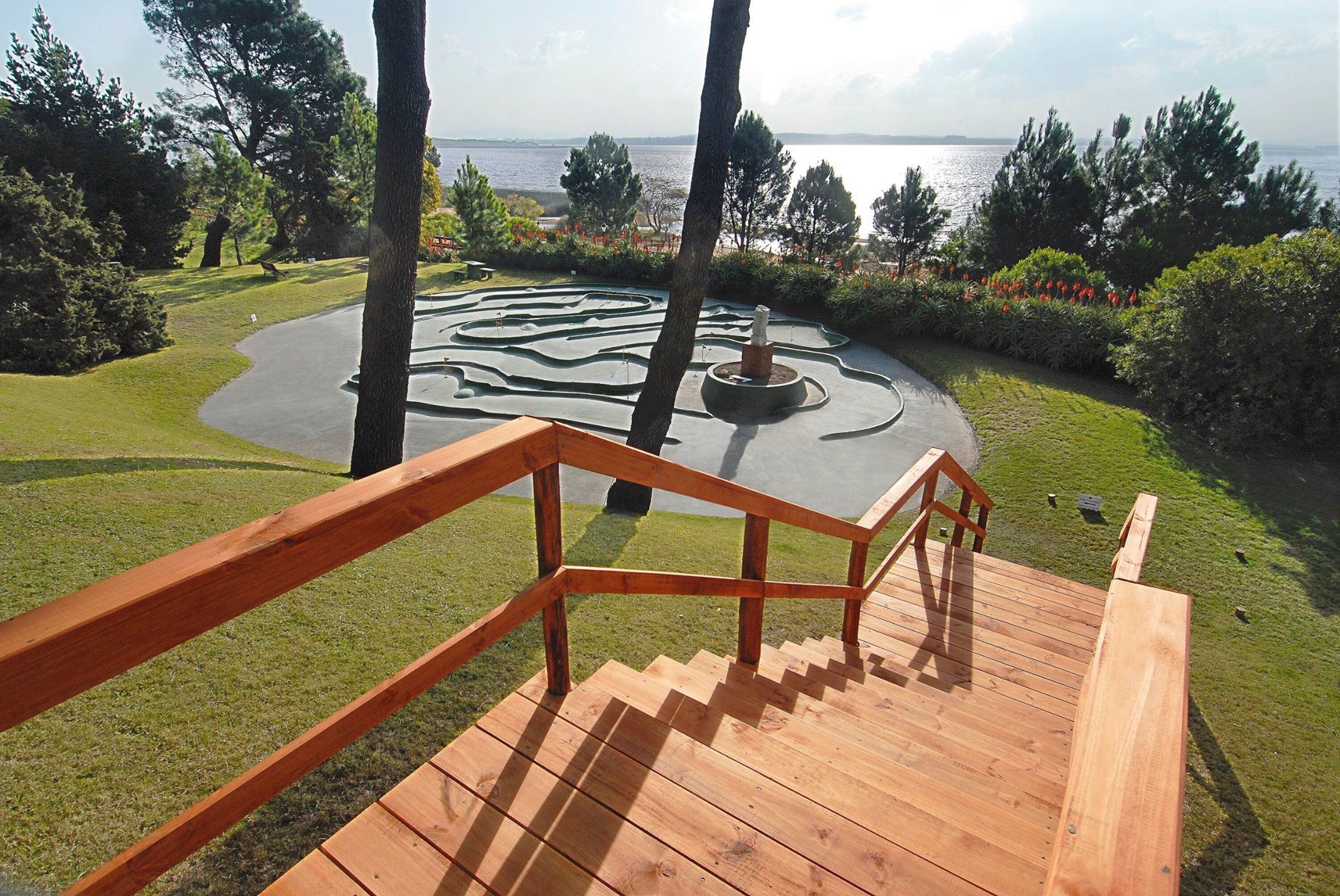 Eine Holztreppe führt zu einem kreisförmigen Garten mit einem Wasserspiel, umgeben von Bäumen und Blick auf das Meer.