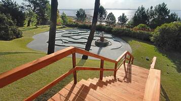 Eine Holztreppe führt zu einem kreisförmigen Garten mit einem Wasserspiel, umgeben von Bäumen und Blick auf das Meer.