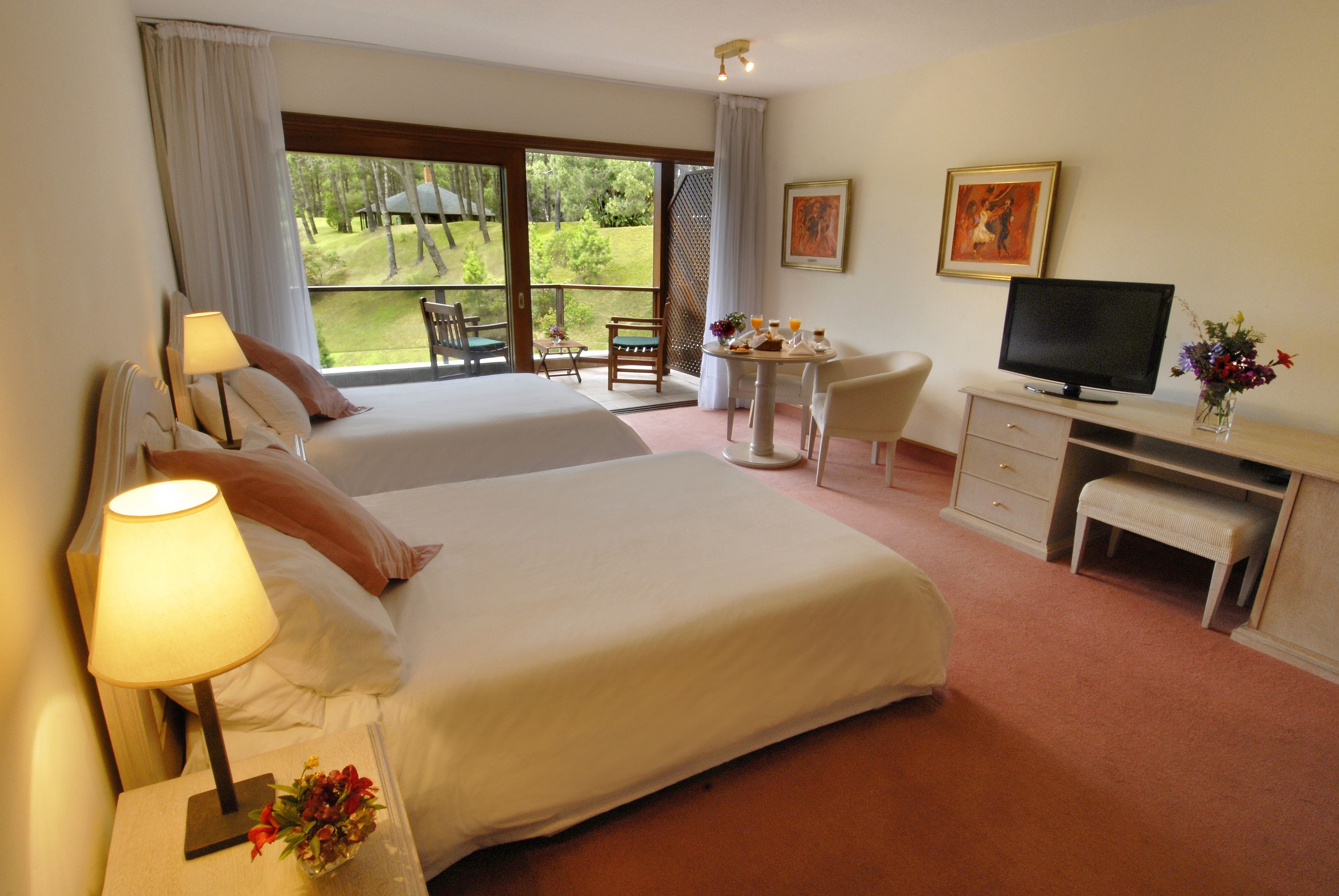 A hotel room with two beds, a desk with a television, a chair, and a small table with a vase of flowers.