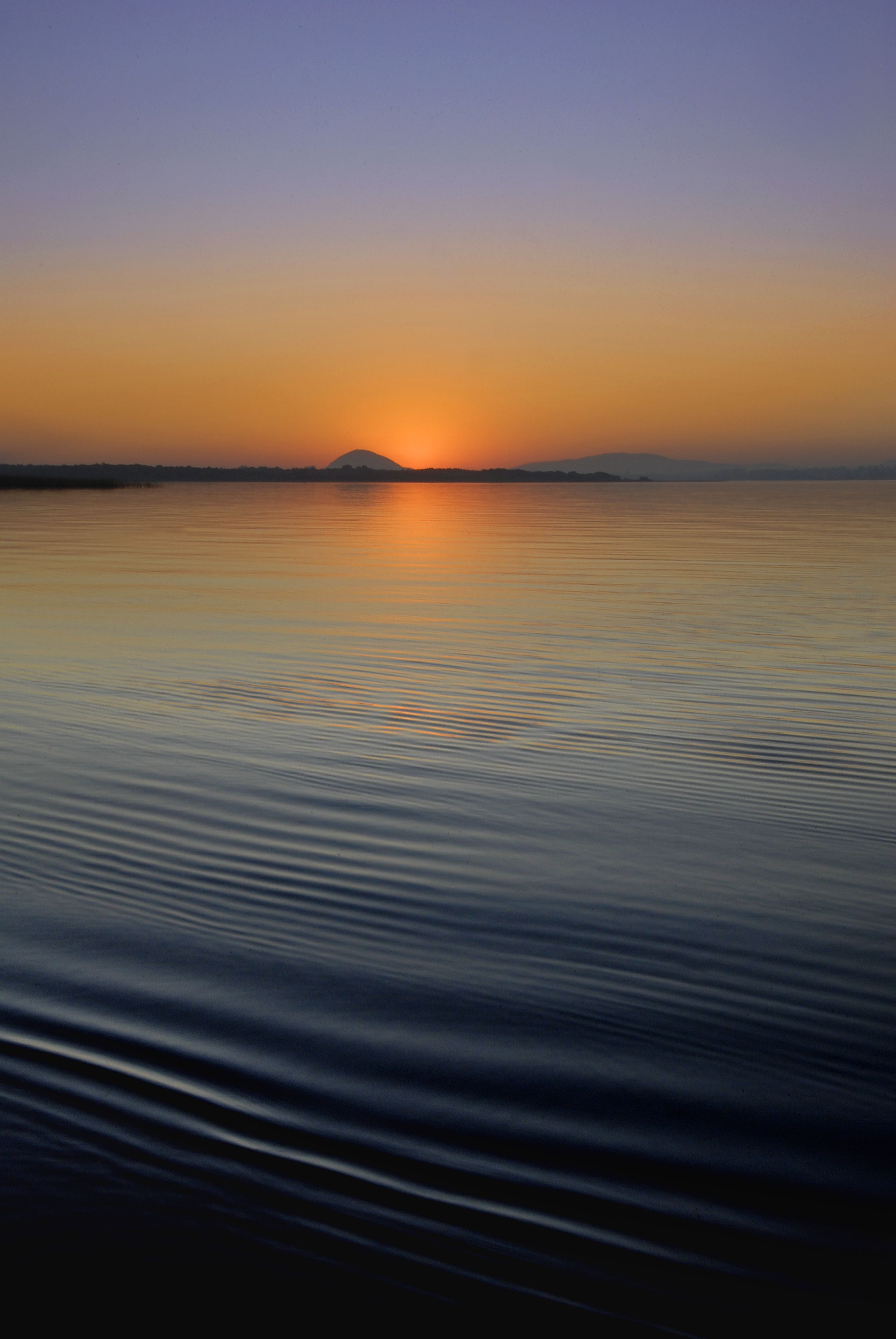 Un cuerpo de agua tranquila que refleja un atardecer con una montaña al fondo.