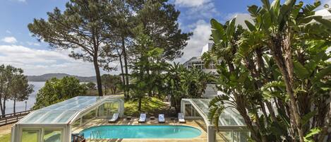 Piscina con tumbonas y estructuras de vidrio, rodeada de árboles y vista al agua.