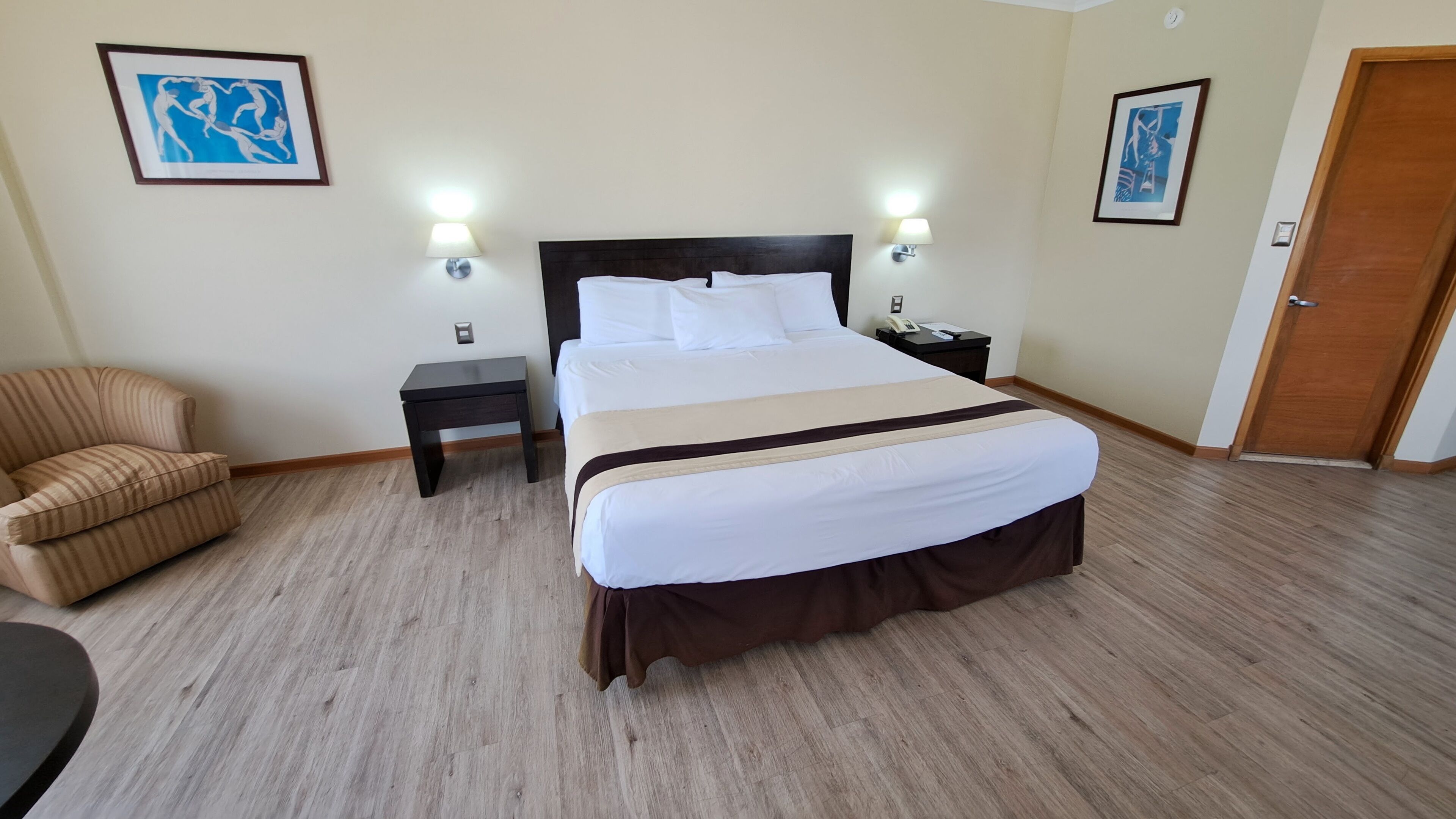 Habitación de hotel con una cama grande, dos mesitas de noche, una silla y dos lámparas fijadas a la pared.