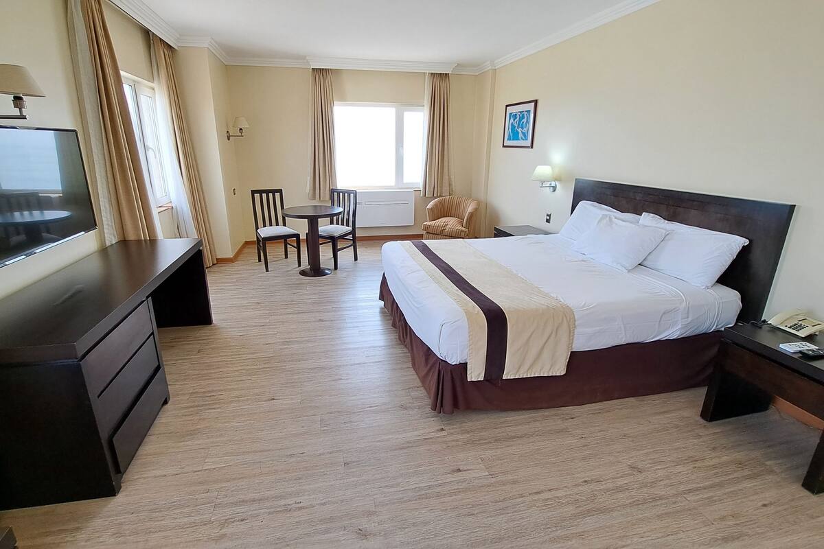 A hotel room with a bed, a desk with a TV, a chair, a table, and a window with curtains.