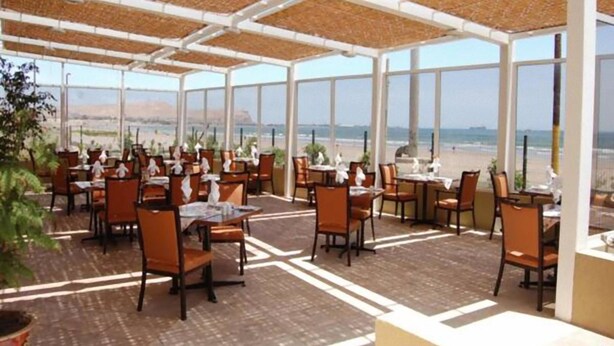 Un espacio de comedor al aire libre con mesas y sillas, ventanas amplias y vista a la playa.