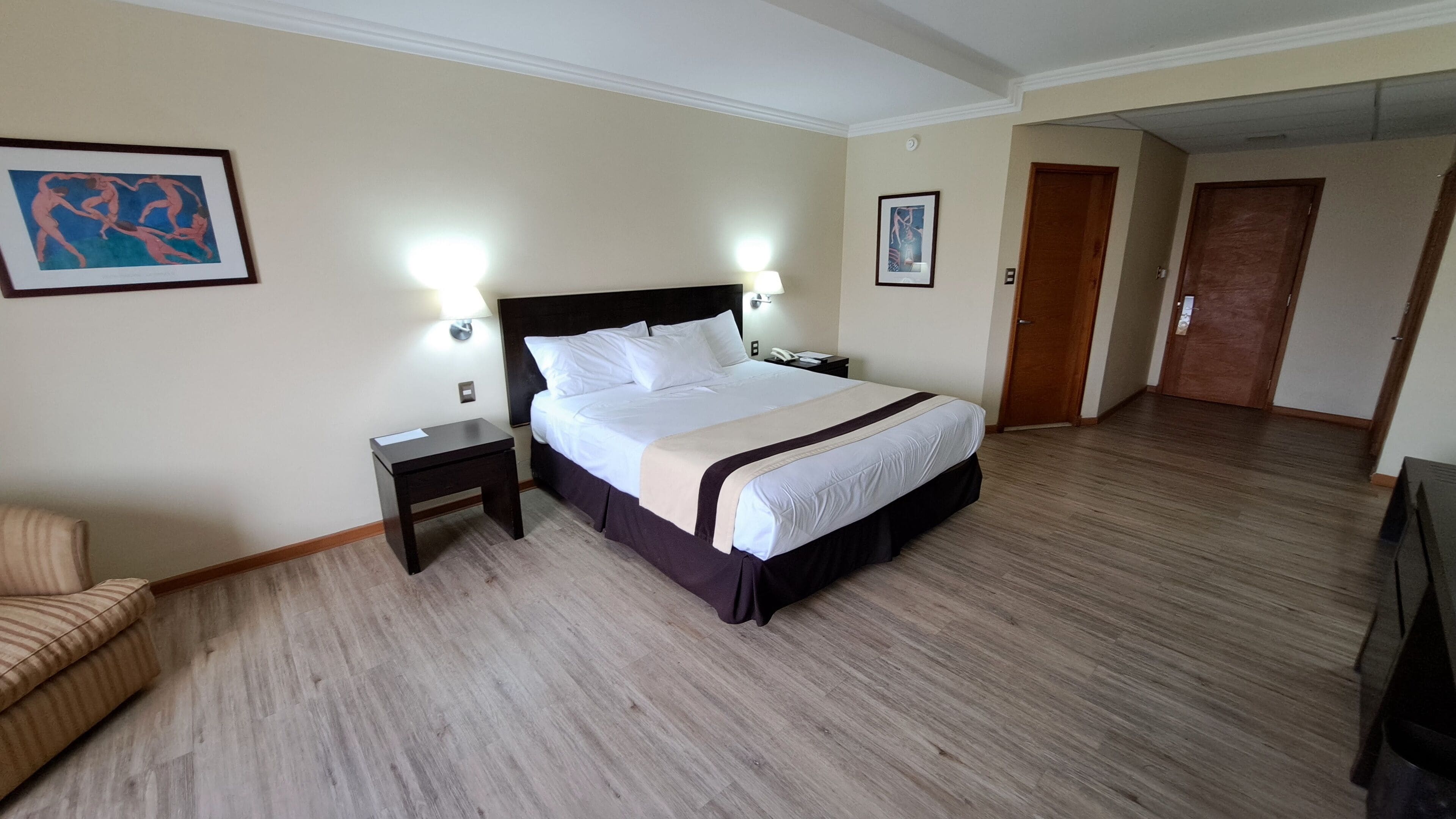 A hotel room with a large bed, two bedside tables, a chair, and two framed artworks on the walls.