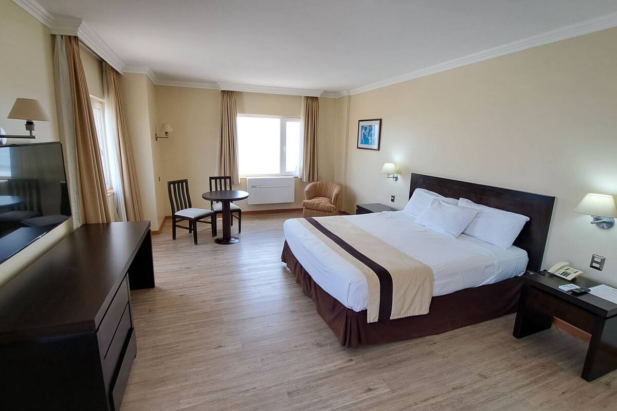 A hotel room with a bed, bedside table, lamp, chair, desk, TV, and a window with curtains.