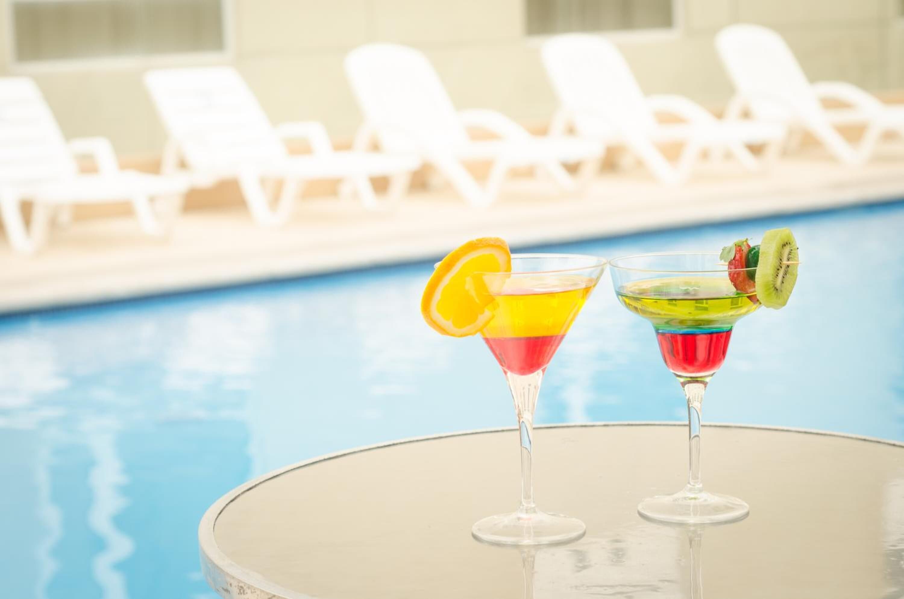 Two cocktails on a table by a poolside with lounge chairs in the background.