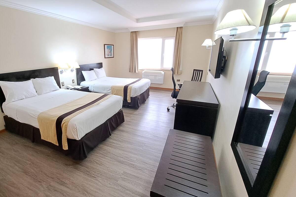 A hotel room with two beds, a desk, a chair, a mirror, and a window with curtains.
