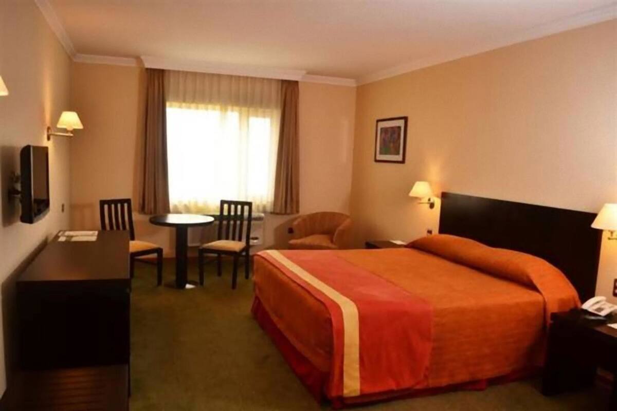 A hotel room with a bed, a desk, a chair, a small table, and a window with curtains.