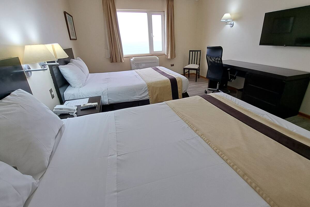 A hotel room with two beds, a desk, a chair, a TV, and a window with curtains.