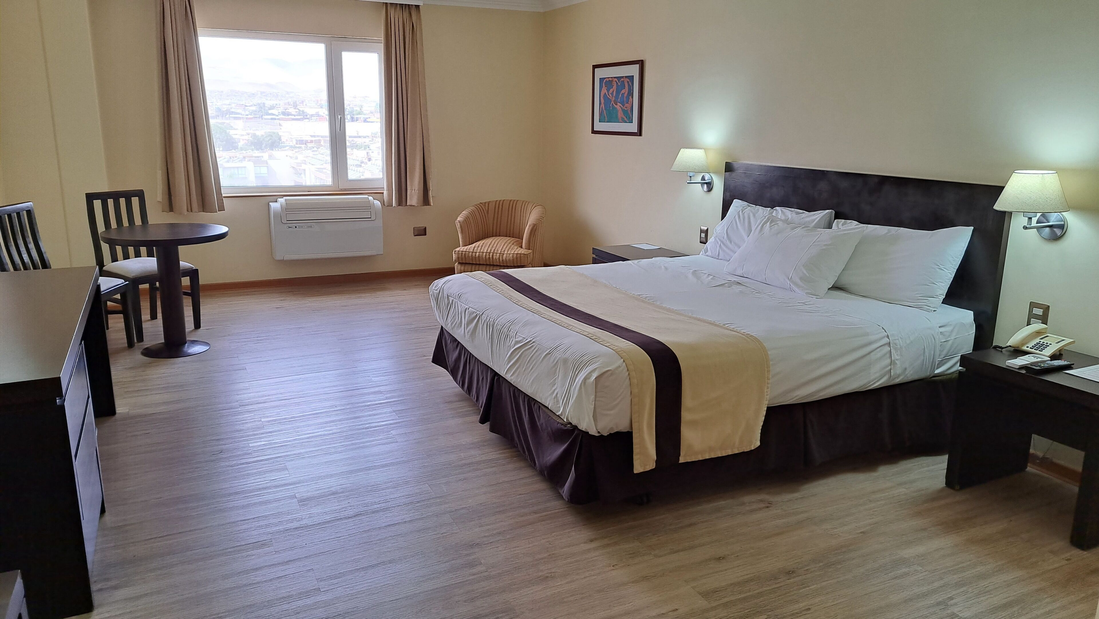 Habitación de hotel con cama, escritorio, silla, ventana con cortinas y lámpara de pared.