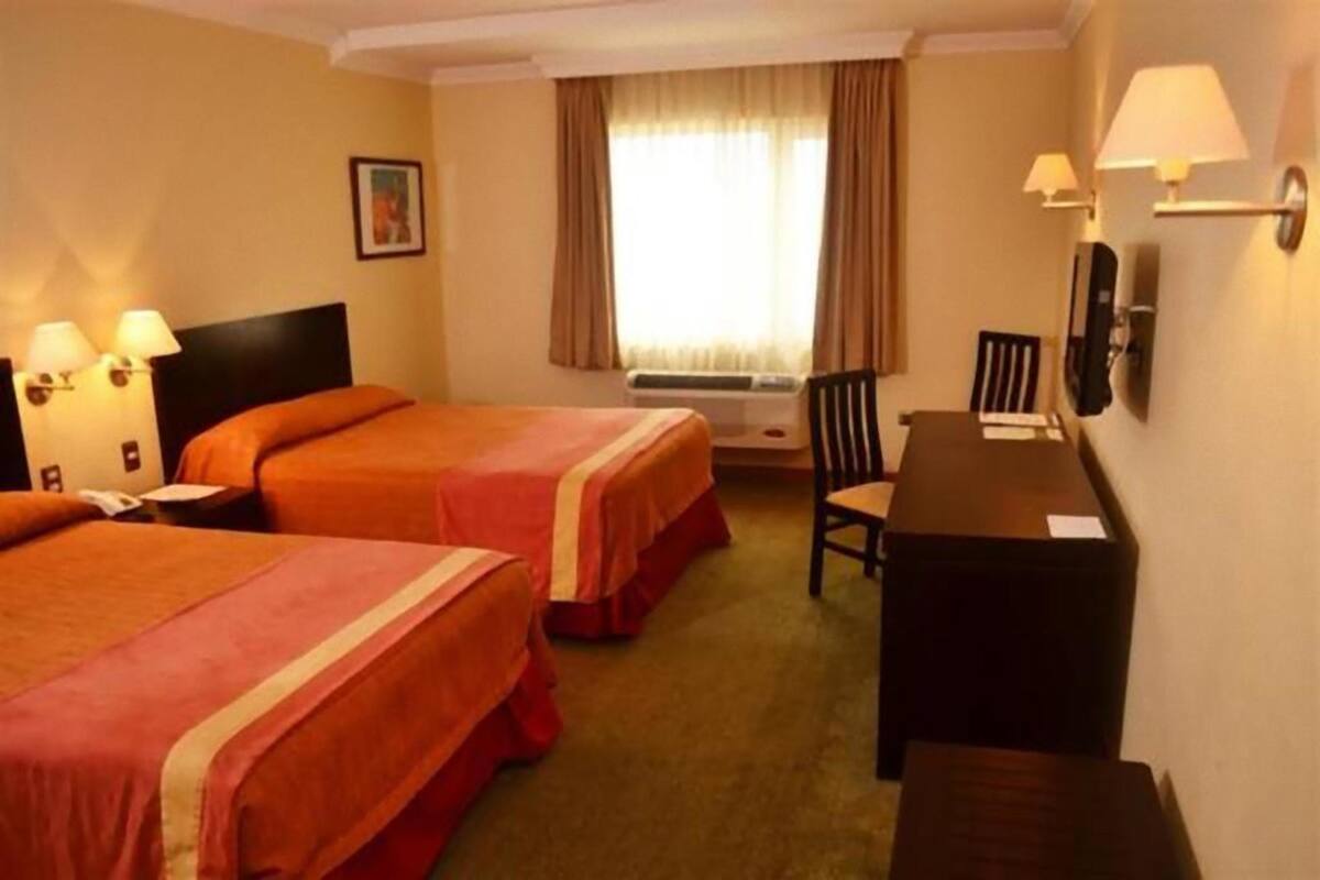 A hotel room with two beds, a desk, a chair, a window with curtains, and a wall-mounted lamp.