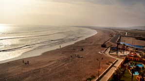 Una playa con vista despejada al océano, un estacionamiento y una pequeña zona de piscina con palmeras.