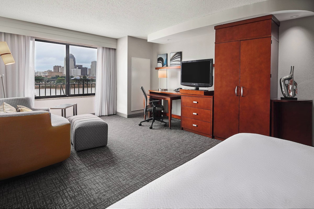 Une chambre d’hôtel avec un grand lit, un bureau avec une chaise, une télévision, une armoire et une vue sur la ville grâce à une grande fenêtre.