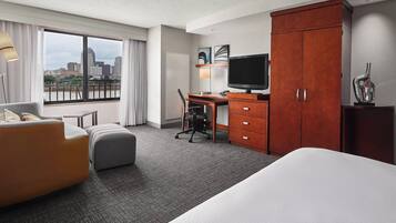 A hotel room with a large bed, a desk with a chair, a television, a wardrobe, and a view of the city through a large window.