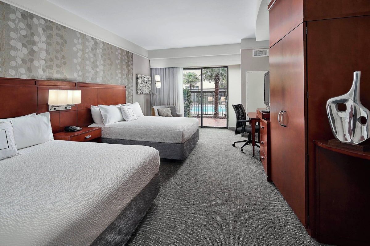 Une chambre d’hôtel avec deux lits, un bureau, une chaise, un grand miroir et une vue sur la piscine, accessible par une porte-fenêtre coulissante.