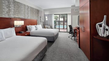 A hotel room with two beds, a desk, a chair, a large mirror, and a view of a pool through a sliding glass door.