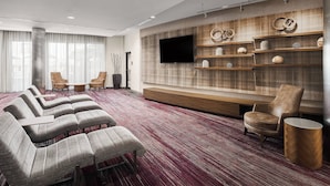 A modern hotel room with a large seating area, a flat-screen TV mounted on a wooden wall, and a decorative shelf with items. - Courtyard by Marriott Shreveport-Bossier/Louisiana Boardwalk (Bossier City)