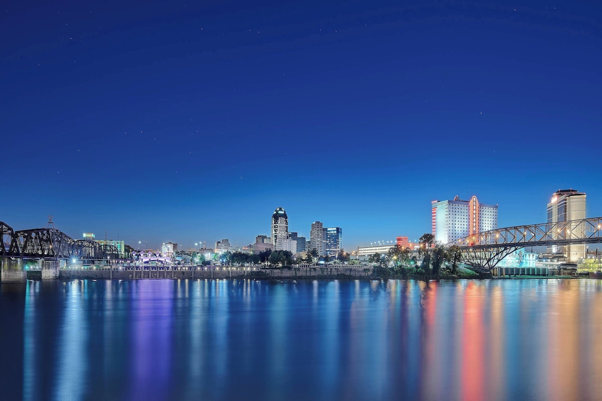 A nighttime cityscape with a river reflecting city lights, a bridge, and high-rise buildings.