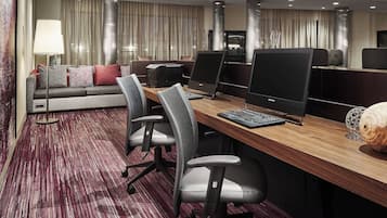A modern hotel room with a long wooden desk, two office chairs, a sofa, and a large flat-screen TV.