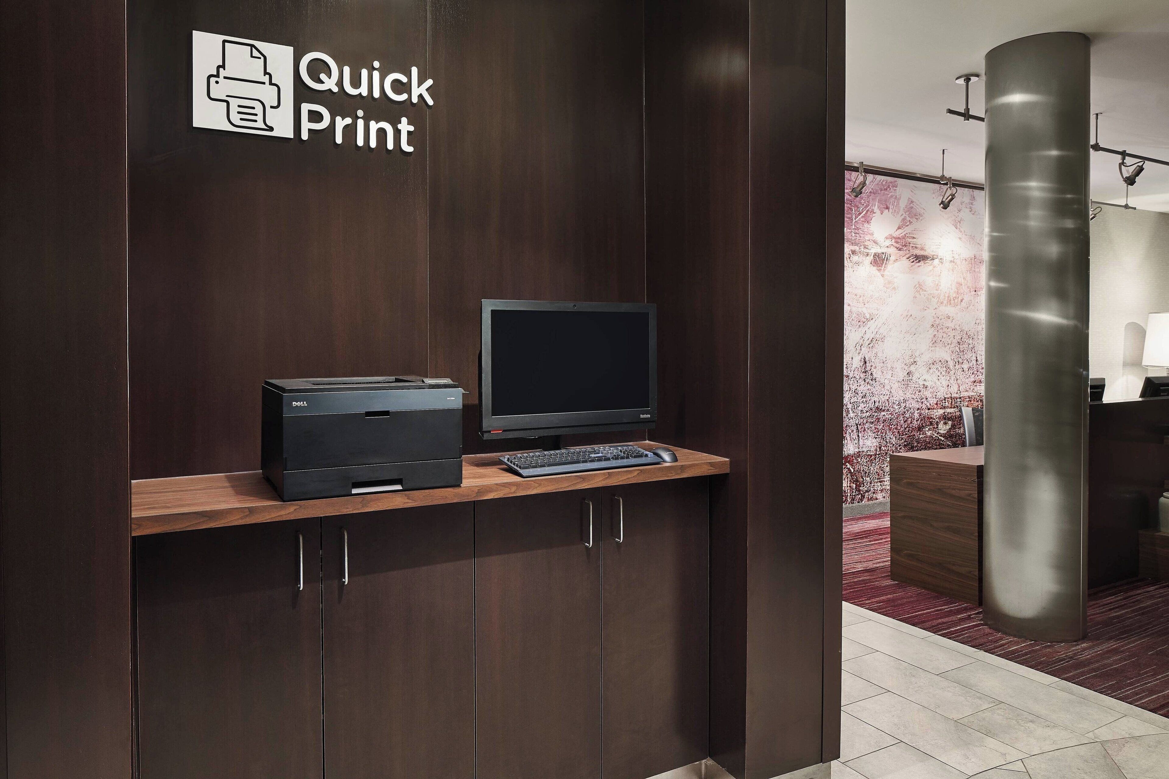 Um balcão de recepção com uma impressora e computador, bancada de madeira e uma placa com a inscrição 'Quick Print'.