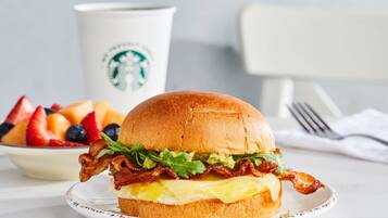 Ein Frühstücks-Sandwich mit Speck, Avocado und Ei auf einem Brötchen, ein Obstteller und eine Starbucks-Tasse im Hintergrund.