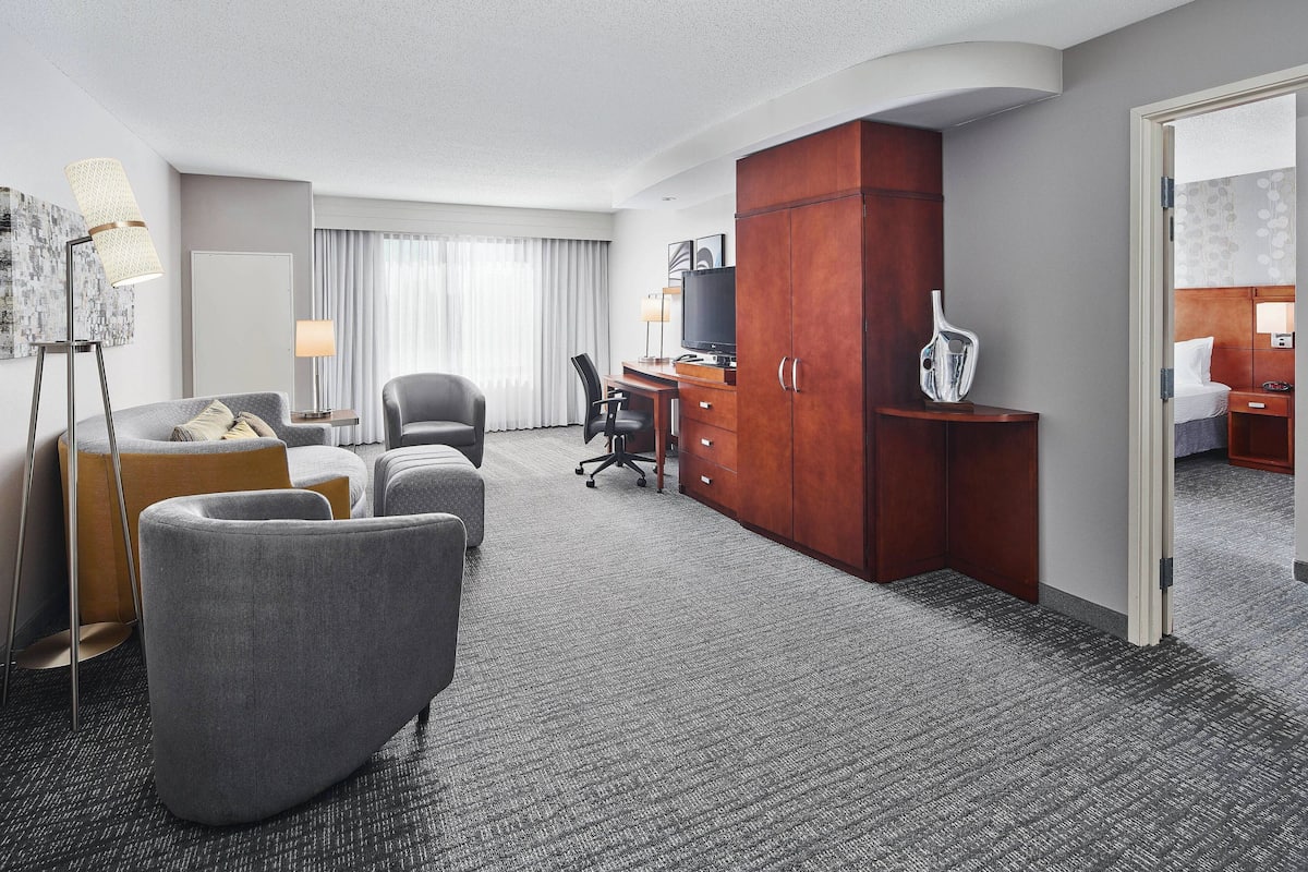 Une chambre d’hôtel avec un tapis gris, un canapé gris, une armoire en bois, un bureau avec un ordinateur, une télévision et un lit avec une couette blanche.