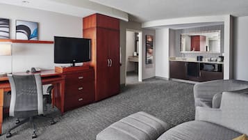 A hotel room with a desk, television, wardrobe, kitchenette, and a grey sofa.